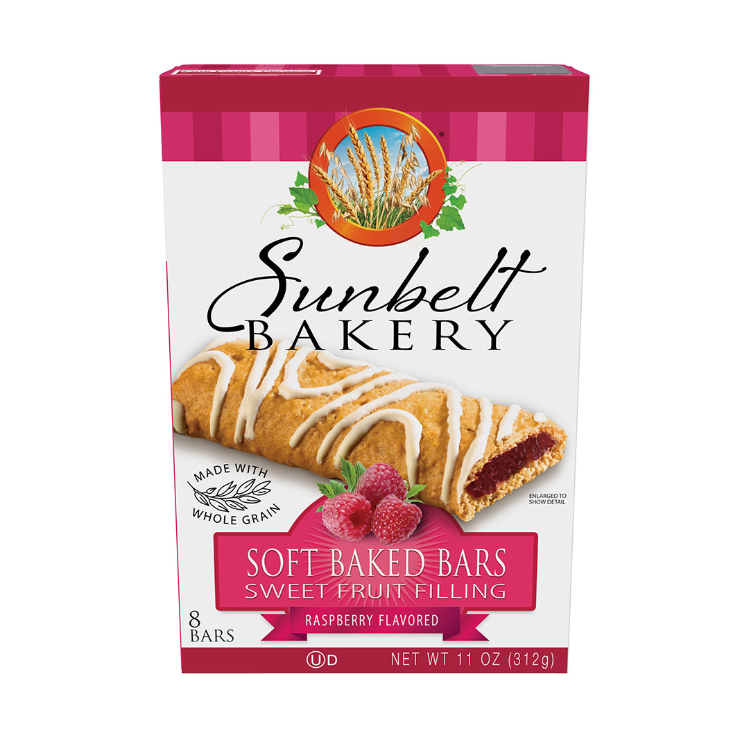 Image of Sunbelt Bakery's Raspberry Flavored Soft Baked Bars packaging. The front of the box features a whole-grain bar with a swirl of white glaze on top, revealing a vibrant raspberry filling. There are also two raspberries next to the bar. The packaging is marked with 'Made with Whole Grain', contains 8 bars, and has a net weight of 11 oz (312g). The box is trimmed with a pink gingham pattern at the top and includes the Sunbelt Bakery logo with a background illustration of golden wheat. 