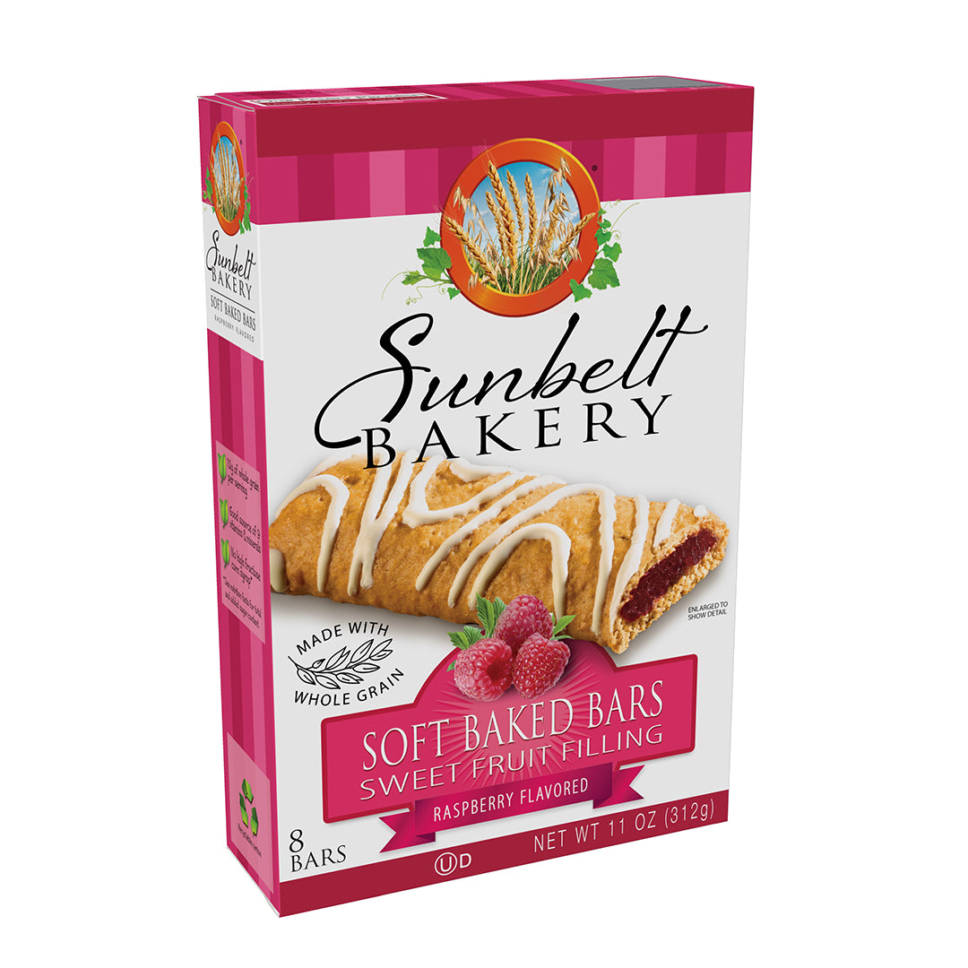 Packaging for Sunbelt Bakery Raspberry Flavored Soft Baked Bars, showing a whole grain bar with white glaze and a raspberry flavored jam filling, accompanied by a cluster of ripe raspberries. The box features a bright pink gingham pattern on the top and side, a 'Made with Whole Grain' claim, and states there are 8 bars with a net weight of 11 oz (312g). The Sunbelt Bakery logo is displayed at the top with an image of wheat stalks in the background.